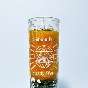 May include: A glass candle with layers of orange, green, and yellow wax. The candle has the words "Trabajo Fijo" and "Steady Work" printed on it, along with illustrations of various professions. The candle is designed to be used for spiritual or ritual purposes.