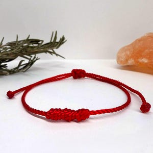 May include: A red string bracelet with a central knot design. The bracelet is made of a thin, woven material and features a sliding knot closure. The background includes a sprig of rosemary and a piece of orange-colored rock salt.
