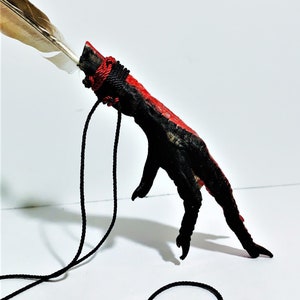May include: A black and red chicken foot with a feather attached to it. The foot is tied with a black cord and a red cord. The foot is hanging from a black cord.