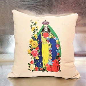 May include: A white pillow with a colorful image of a skeleton wearing a crown and a blue and green robe. The skeleton is surrounded by flowers, including sunflowers, roses, and blue flowers. The pillow is a decorative item for a home.