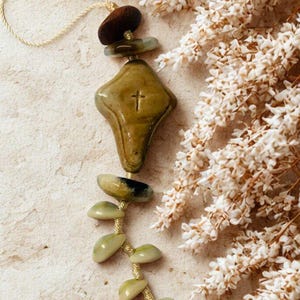 May include: A religious pendant with a cross design. The olive green pendant has a small cross etched into the centre. It is strung with a gold cord and decorated with natural stone beads.
