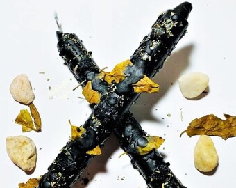May include: Two black candles with gold flecks and dried leaves scattered around them on a white background.