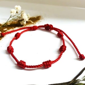 Peut inclure: Un bracelet rouge en cordon avec des perles nouées. Le bracelet est sur une surface blanche avec des fleurs et des feuilles séchées.