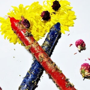 May include: Two crossed ritual candles, one red and one blue, are adorned with dried herbs and gold flecks. The candles are surrounded by yellow chrysanthemums and small, purple globe amaranth flowers, creating a vibrant, spiritual aesthetic.