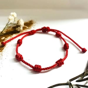 May include: A red string bracelet with seven decorative knots. The bracelet is made of a twisted red cord and is displayed on a white surface. The bracelet is a simple, yet elegant piece of jewelry.