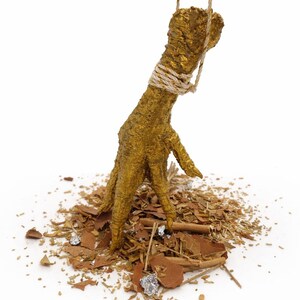 May include: A golden chicken foot sculpture, suspended by a rope, stands on a bed of brown and silver fragments. The foot is intricately detailed, with a textured surface and a rope wrapped around the ankle.