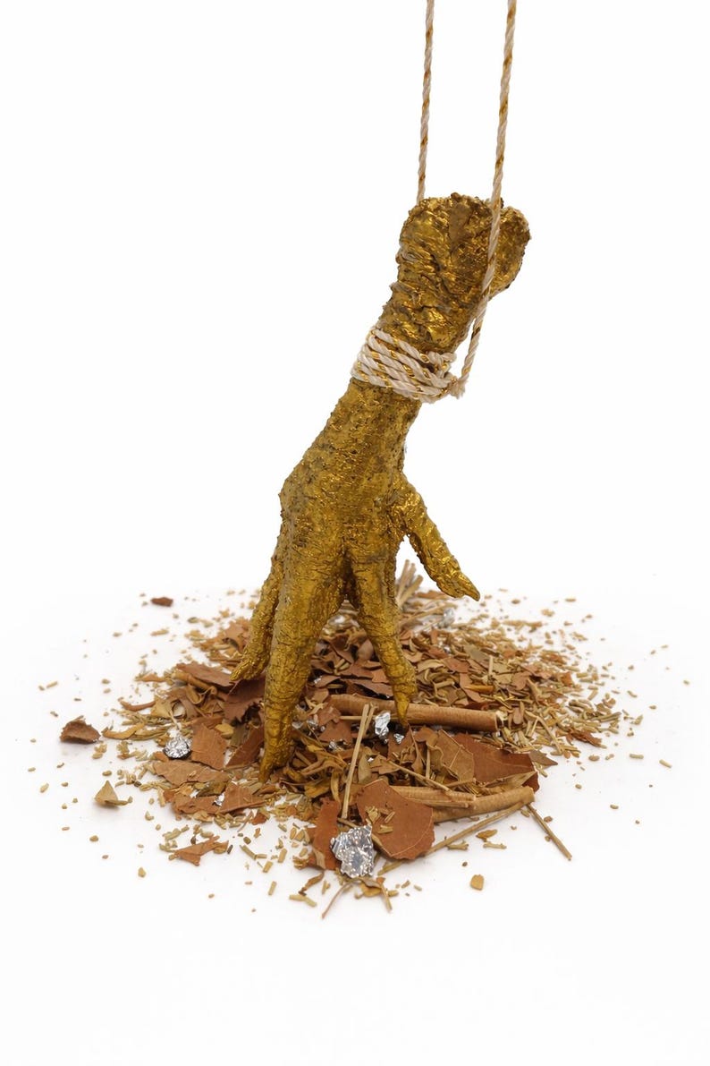 May include: A golden chicken foot sculpture, suspended by a rope, stands on a bed of brown and silver fragments. The foot is intricately detailed, with a textured surface and a rope wrapped around the ankle.