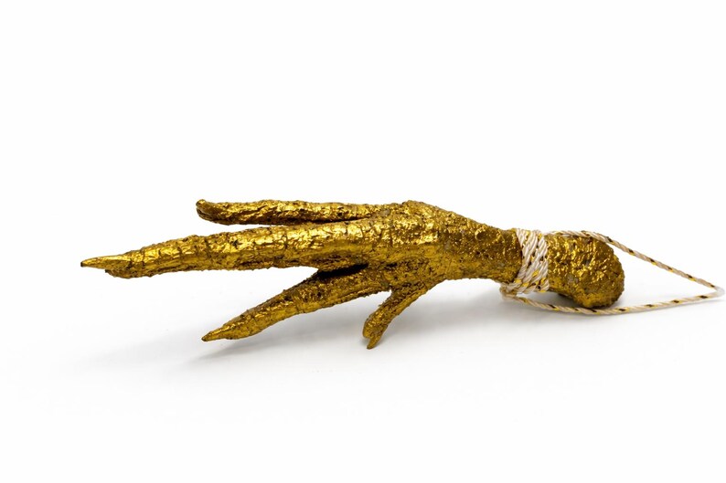 May include: A gold-colored, textured decorative object shaped like a bird's foot. The foot has three long toes and a textured surface. A white and gold string is wrapped around the base of the foot.