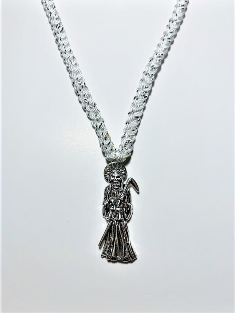 May include: A white braided cord necklace with a silver pendant featuring a skeletal figure holding a scythe. The pendant is a depiction of Santa Muerte, a folk saint associated with death and protection.