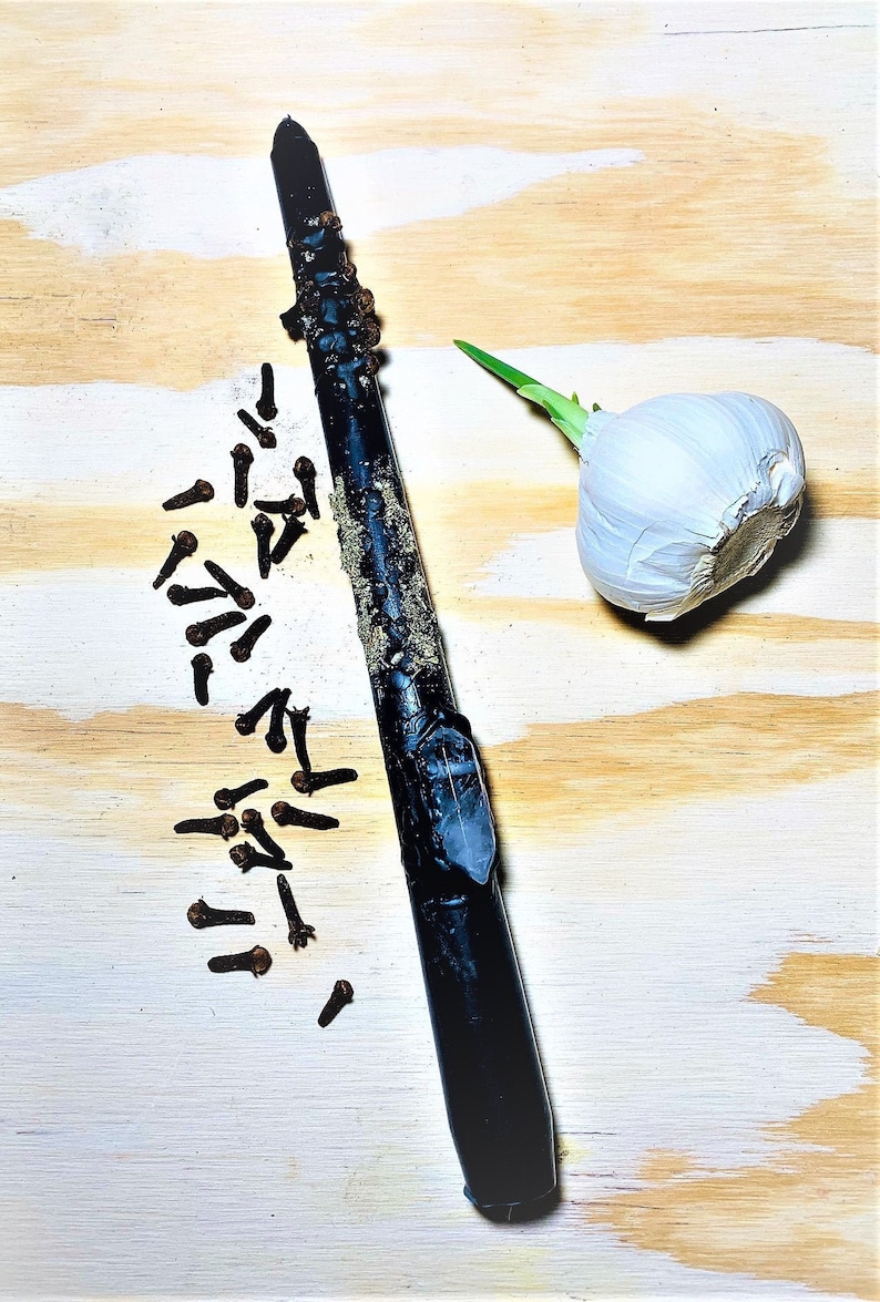 Peut inclure: Une bougie noire avec des clous de girofle &eacute;parpill&eacute;s autour et un bulbe d'ail blanc avec une tige verte sur une surface en bois.