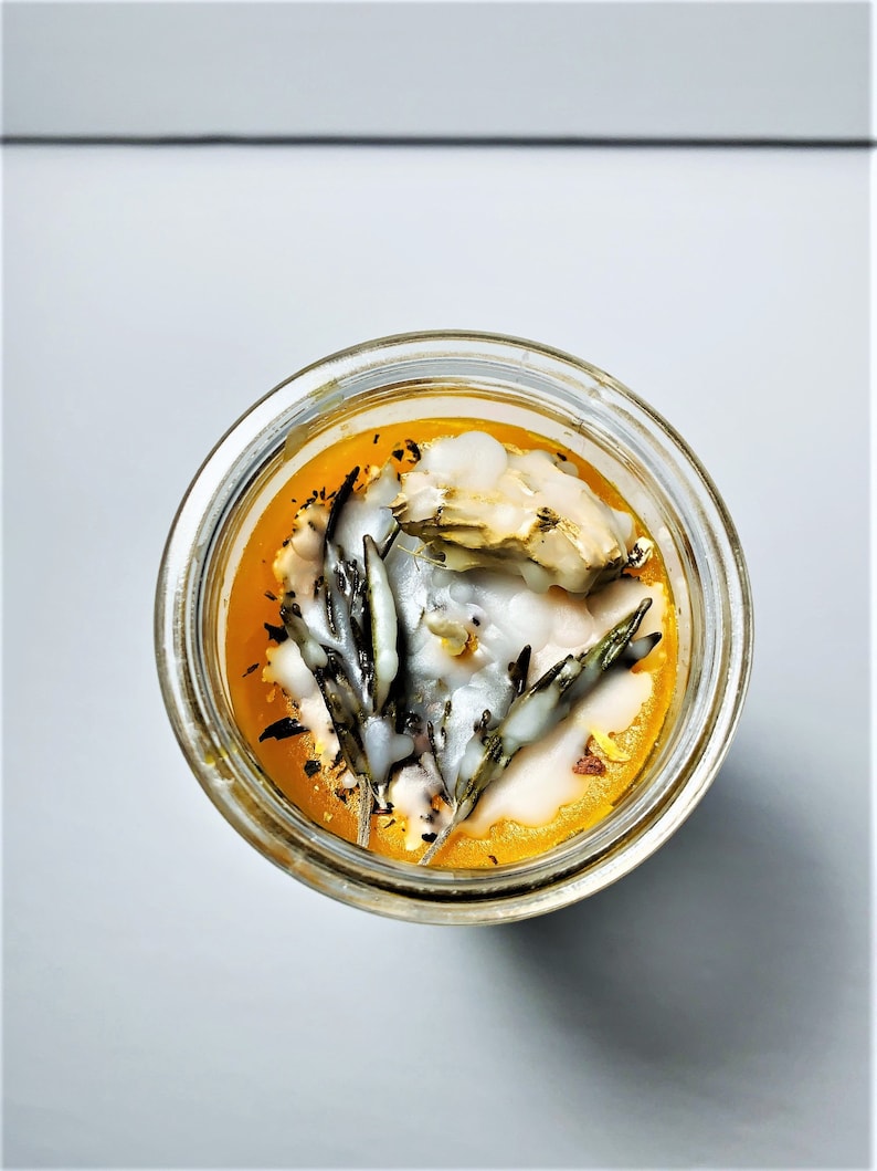 May include: Overhead shot of a clear glass jar filled with a vibrant orange liquid, topped with white wax and sprigs of rosemary. The jar's contents are illuminated, creating a warm, inviting glow. The overall aesthetic suggests a handmade candle or cosmetic product.