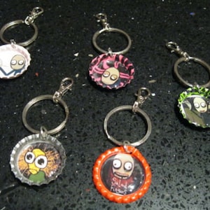 May include: Six keychains with different designs featuring cartoon characters. Each keychain is made from a bottle cap with a silver metal ring. The characters include a man with a pink shirt, a man with a green shirt, a man with a blue shirt, a man with a red shirt, a man with a yellow shirt, and a man with a black shirt.