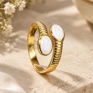May include: A gold-toned bypass ring featuring a ribbed, textured band that tapers into two open ends. Each end is capped with a smooth, oval-shaped white stone inlay, creating a balanced design with a polished metallic finish throughout the band.