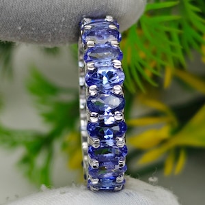 May include: A silver ring featuring a continuous row of oval-cut, deep blue gemstones. The ring's design showcases the gemstones set closely together, creating a band of vibrant color. The ring is held against a backdrop of green foliage.