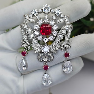 Peut inclure: Broche argentée ornée d'une grande pierre rouge carrée au centre, entourée de petits cristaux transparents. La broche présente un motif floral avec des pierres transparentes en forme de poire et de petites pierres rouges.