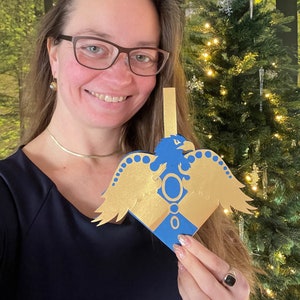 May include: A blue and gold Christmas ornament shaped like a heart with a golden eagle design. The ornament is hanging from a string and has a gold circle with a white dot in the center.