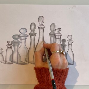 May include: A hand draws a series of glass bottles in pencil on white paper. The bottles are arranged in a row, with the tallest bottle in the center. The hand is wearing a red sweater and a ring.