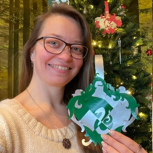 May include: A woman holds a green and silver Christmas ornament featuring the Hogwarts house crest for Slytherin. The crest has a snake and the letters "S" and "L" intertwined.