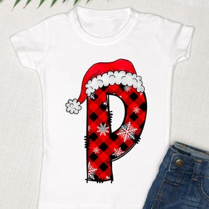 May include: White t-shirt with a red and black plaid letter "P" wearing a Santa hat. The letter is decorated with white snowflakes.