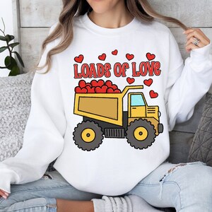May include: White sweatshirt with a yellow dump truck filled with red hearts. The truck has the words "Loads of Love" printed above it.