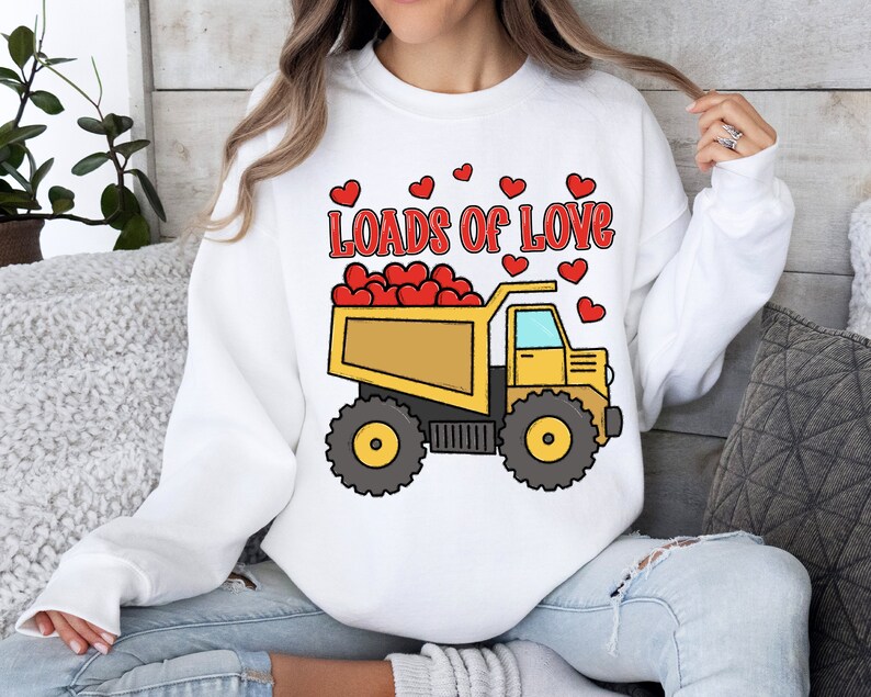 May include: White sweatshirt with a yellow dump truck filled with red hearts. The truck has the words "Loads of Love" printed above it.