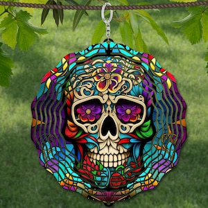 May include: A colorful metal wind spinner featuring a detailed sugar skull design with flowers and swirls. The skull is white with black eyes and a pink tongue. The background is a mix of blue, green, purple, and yellow.