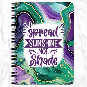 May include: Spiral-bound notebook with a vibrant cover featuring a marbled design in shades of green, purple, and gold. The cover includes the text "spread SUNSHINE NOT Shade" in white and purple.
