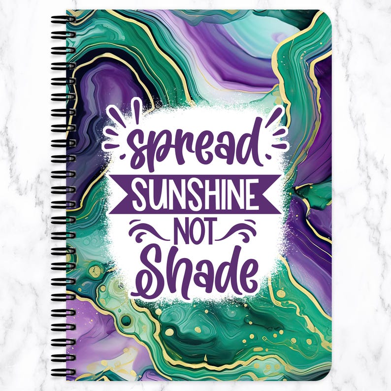 May include: Spiral-bound notebook with a vibrant cover featuring a marbled design in shades of green, purple, and gold. The cover includes the text "spread SUNSHINE NOT Shade" in white and purple.