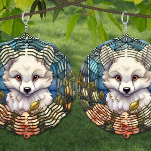 May include: A pair of stained glass earrings featuring a white fox with orange eyes, surrounded by roses and a blue background. The earrings are suspended from silver hooks.