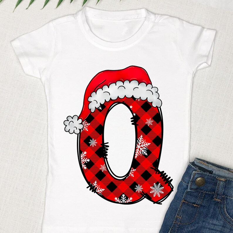 May include: White t-shirt with a red and black plaid letter Q wearing a Santa hat. The letter Q is decorated with white snowflakes.