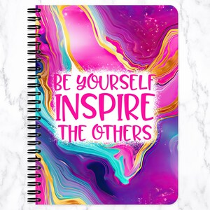 May include: A spiral-bound notebook with a vibrant, abstract design in shades of pink, purple, gold, and teal. The cover features the text "BE YOURSELF INSPIRE THE OTHERS" in a bold, pink font, set against a white background.