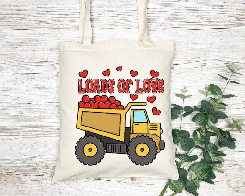 May include: A yellow and black cartoon dump truck filled with red hearts. The truck has the words "Loads of Love" written on the side. The truck is on a white tote bag with a brown handle.