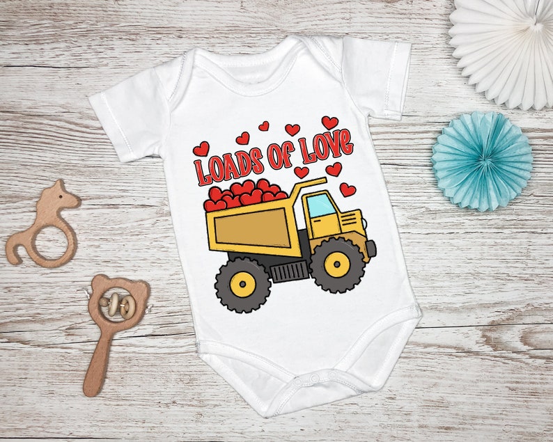 May include: White baby onesie with a yellow dump truck filled with red hearts. The text "Loads of Love" is above the truck.