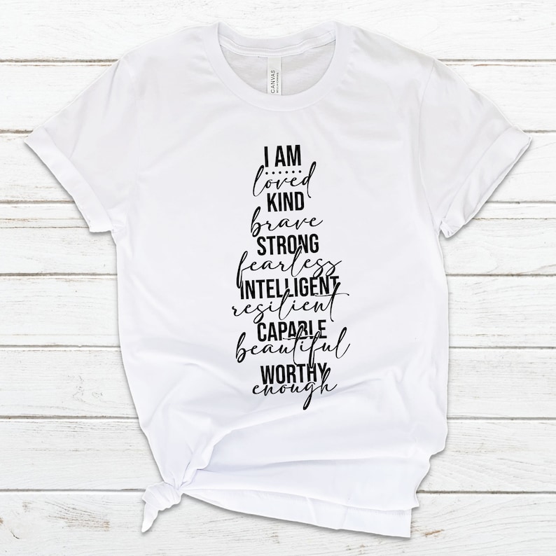 May include: White t-shirt with black text that reads "I am loved kind brave strong fearless intelligent resilient capable beautiful worthy enough".
