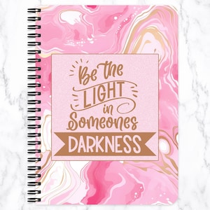 May include: A spiral-bound notebook with a pink marble design and gold accents. The cover features the text "Be the LIGHT in Someones DARKNESS" in a brown font. The notebook has a white marble background.