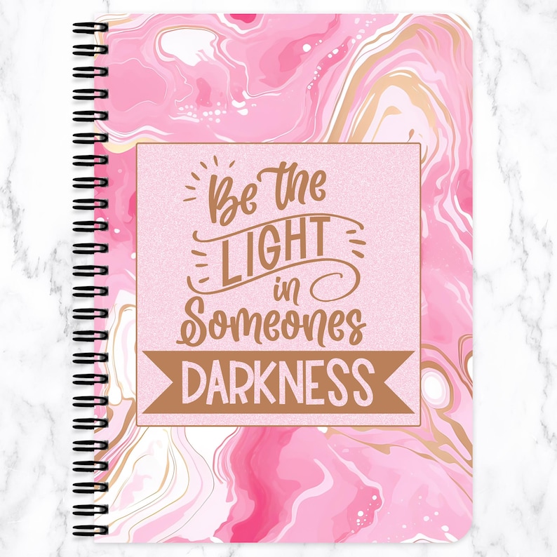 May include: A spiral-bound notebook with a pink marble design and gold accents. The cover features the text "Be the LIGHT in Someones DARKNESS" in a brown font. The notebook has a white marble background.