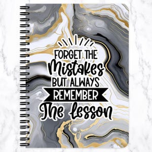 May include: Spiral-bound notebook with a cover featuring a marbled design in shades of gray, black, and gold. The notebook cover has the text "FORGET THE Mistakes BUT ALWAYS REMEMBER The lesson" in a bold, handwritten style.