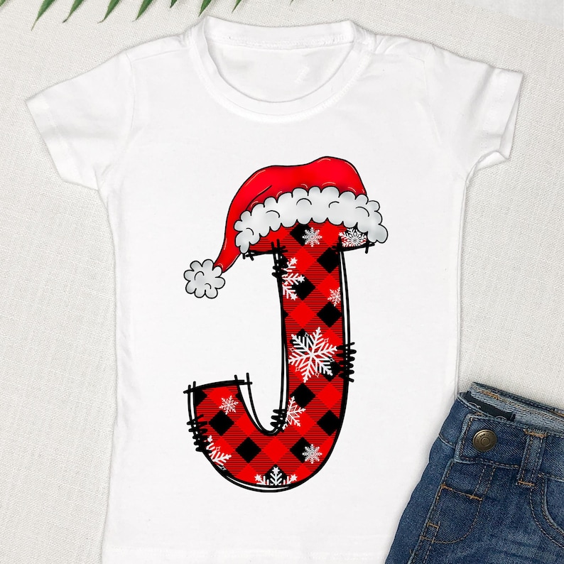 May include: White t-shirt with a red and black plaid letter J with a Santa hat on top. The letter is decorated with white snowflakes.