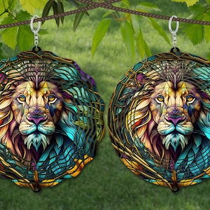 May include: A pair of colorful, circular earrings featuring a detailed, geometric lion design with a mandala-like background. The lion's mane is a vibrant mix of blue, green, and yellow, while its face is a blend of purple, orange, and gold. The earrings are suspended from silver hooks.