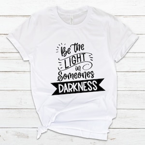 May include: White t-shirt with black text that reads "Be the light in someone's darkness".