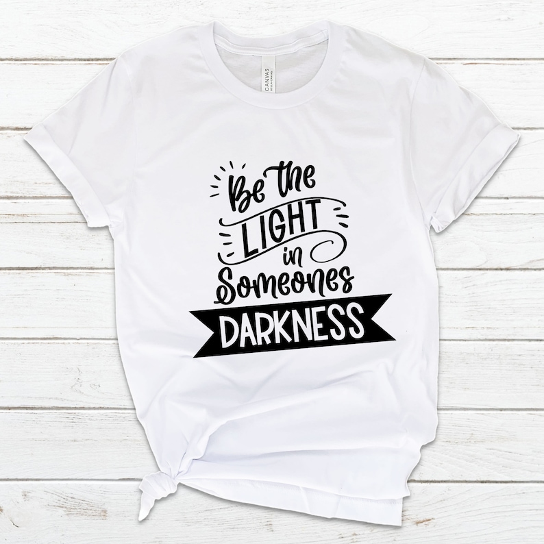 May include: White t-shirt with black text that reads "Be the light in someone's darkness".