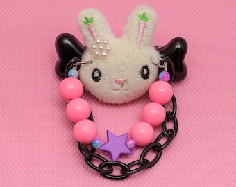 May include: A white plush bunny with embroidered carrot details, black wings, and pink and purple beads attached to a black chain.