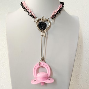 Può includere: Una collana unica con una catena nera e rosa, un ciondolo a forma di cuore argentato con un centro a forma di cuore nero e una lunga spilla argentata. Un ciuccio rosa è attaccato in basso. La collana è esposta su un manichino bianco.