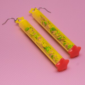 May include: A pair of yellow tube earrings with red star-shaped tops. The tubes are filled with small green and yellow flowers.