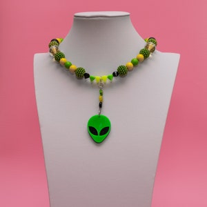 May include: A green alien face charm hangs from a necklace made of yellow, green, and black beads.