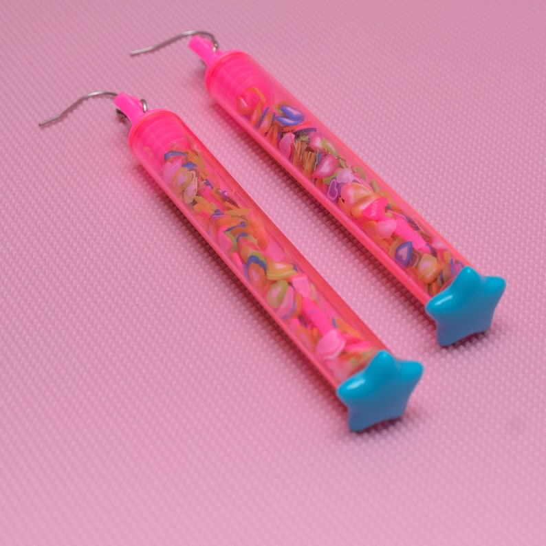 May include: A pair of pink earrings with a clear tube filled with colorful confetti and a blue star charm at the end.