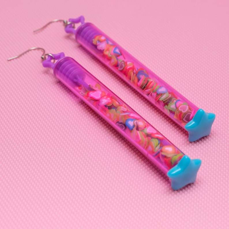 May include: A pair of long, purple and blue earrings with a star-shaped charm at the end. The earrings are filled with colorful hearts.