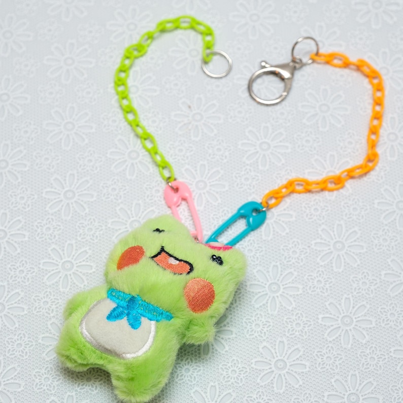 Peut inclure: Un jouet en peluche de grenouille verte avec un n&oelig;ud papillon bleu est attach&eacute; &agrave; une cha&icirc;ne en plastique multicolore avec des &eacute;pingles de s&ucirc;ret&eacute;. La cha&icirc;ne est attach&eacute;e &agrave; un porte-cl&eacute;s argent&eacute;.