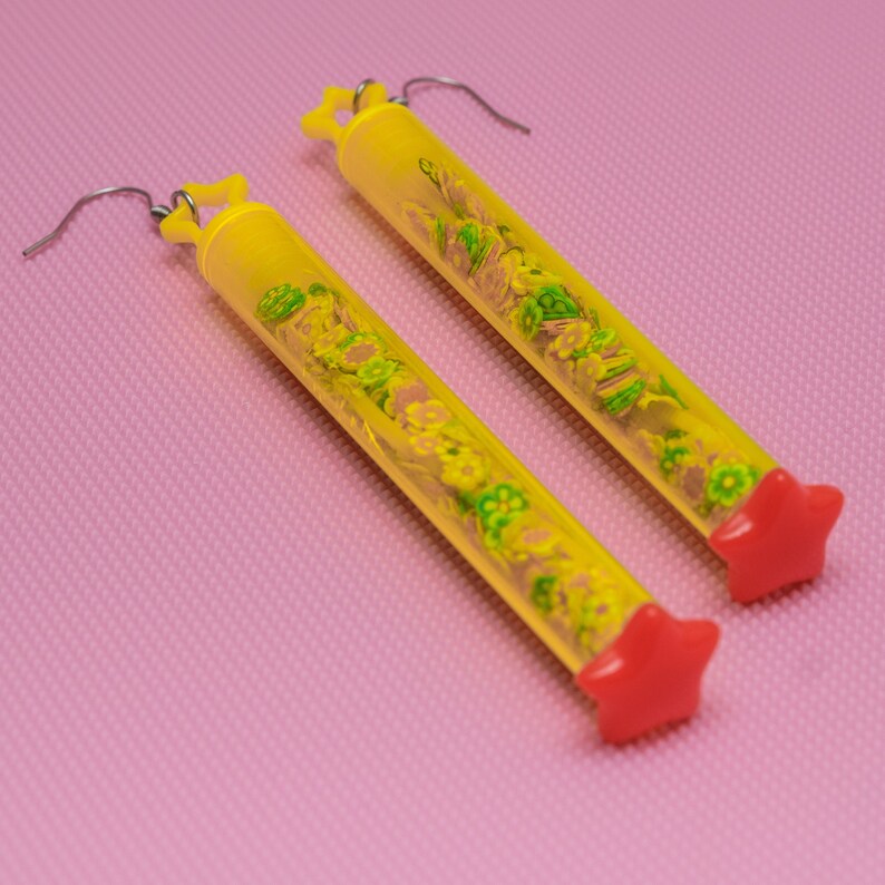May include: A pair of yellow tube earrings with red star-shaped tops. The tubes are filled with small green and yellow flowers.