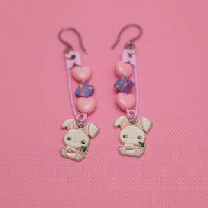 Peut inclure: Une paire de boucles d'oreilles pendantes de couleur rose, violette et blanche. Chaque boucle d'oreille est orn&eacute;e d'un petit charme de lapin blanc, de c&oelig;urs roses et d'un charme carr&eacute; violet.